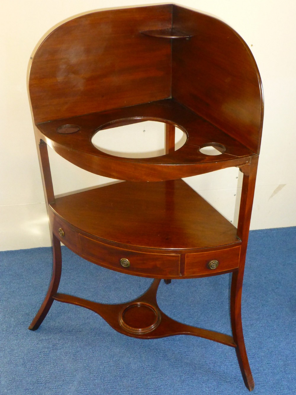 An 18th-century Georgian mahogany corner washstand featuring a curved splashback, a top tier with basin aperture, a lower shelf with a drawer, and curved legs.
