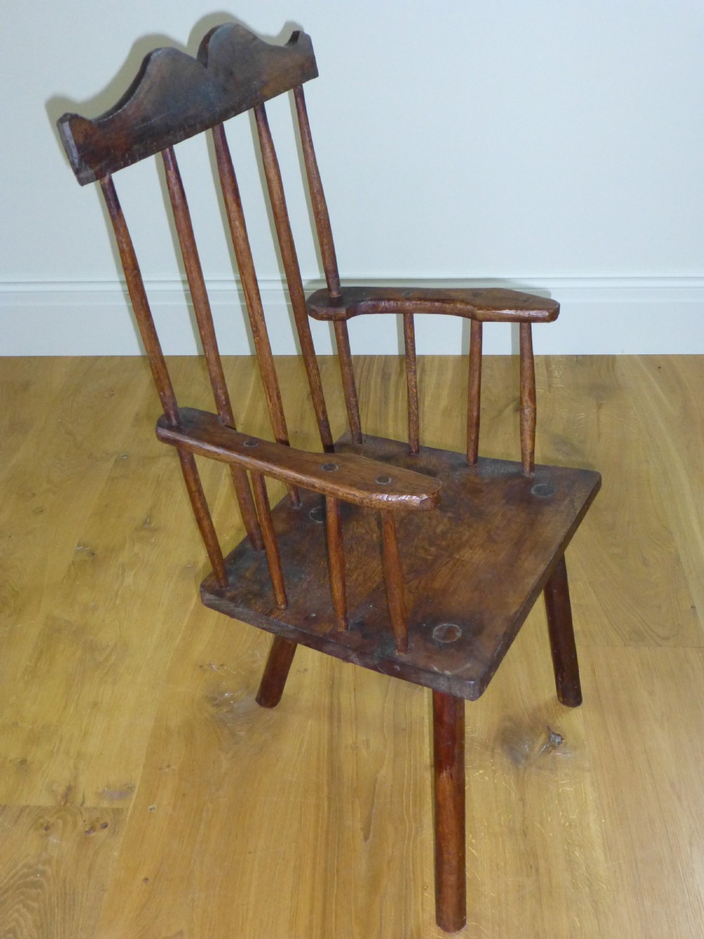 A primitive dark brown wooden Welsh stick armchair featuring a comb back crest rail, hand-shaped spindles, flat armrests, and a solid slab seat on splayed legs.