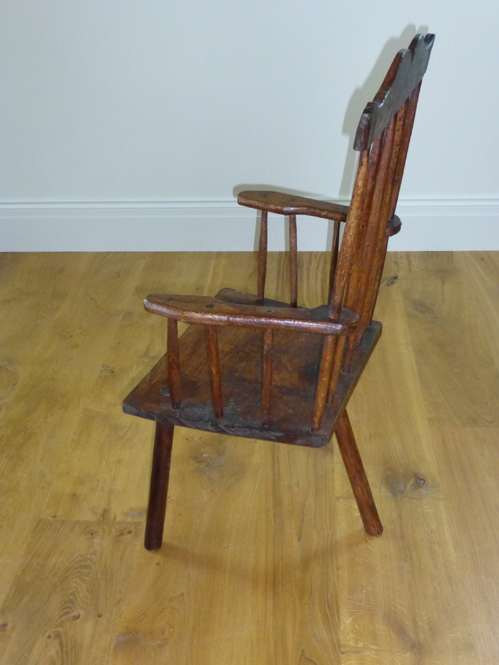 A primitive dark brown wooden Welsh stick armchair featuring a comb back crest rail, hand-shaped spindles, flat armrests, and a solid slab seat on splayed legs. View II