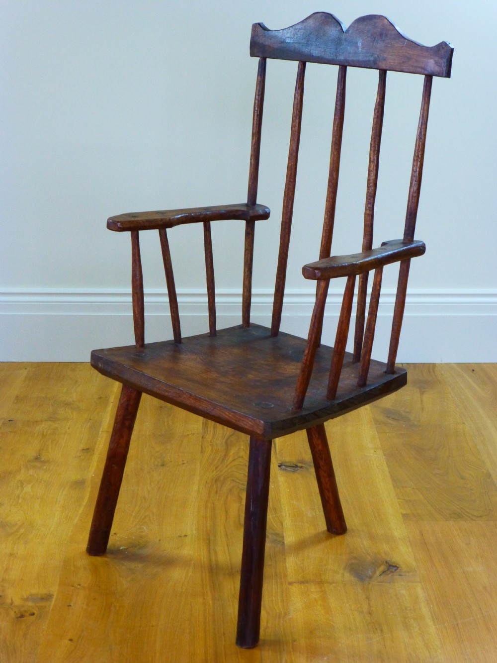 A primitive dark brown wooden Welsh stick armchair featuring a comb back crest rail, hand-shaped spindles, flat armrests, and a solid slab seat on splayed legs. View V