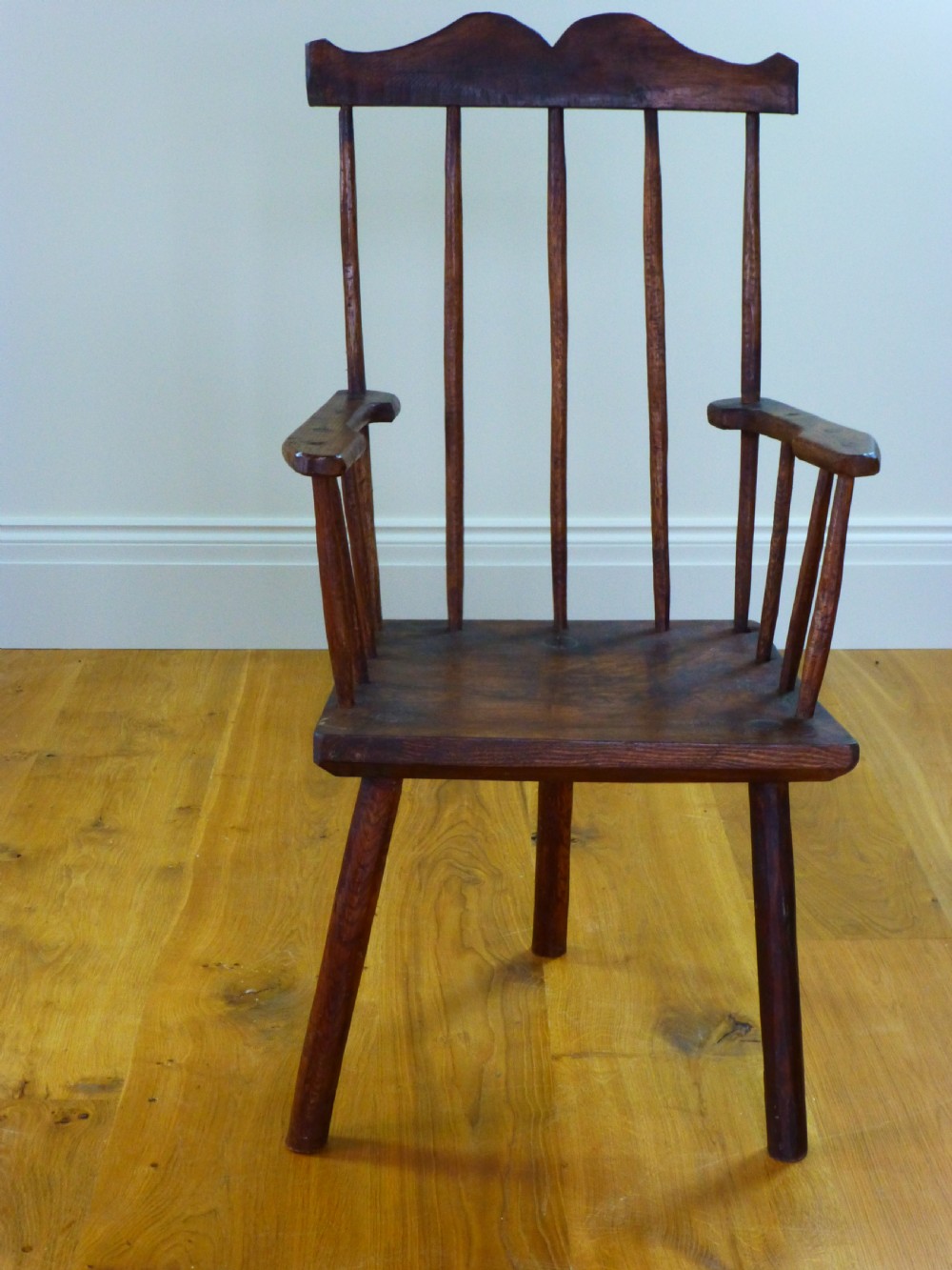 A primitive dark brown wooden Welsh stick armchair featuring a comb back crest rail, hand-shaped spindles, flat armrests, and a solid slab seat on splayed legs. View VI
