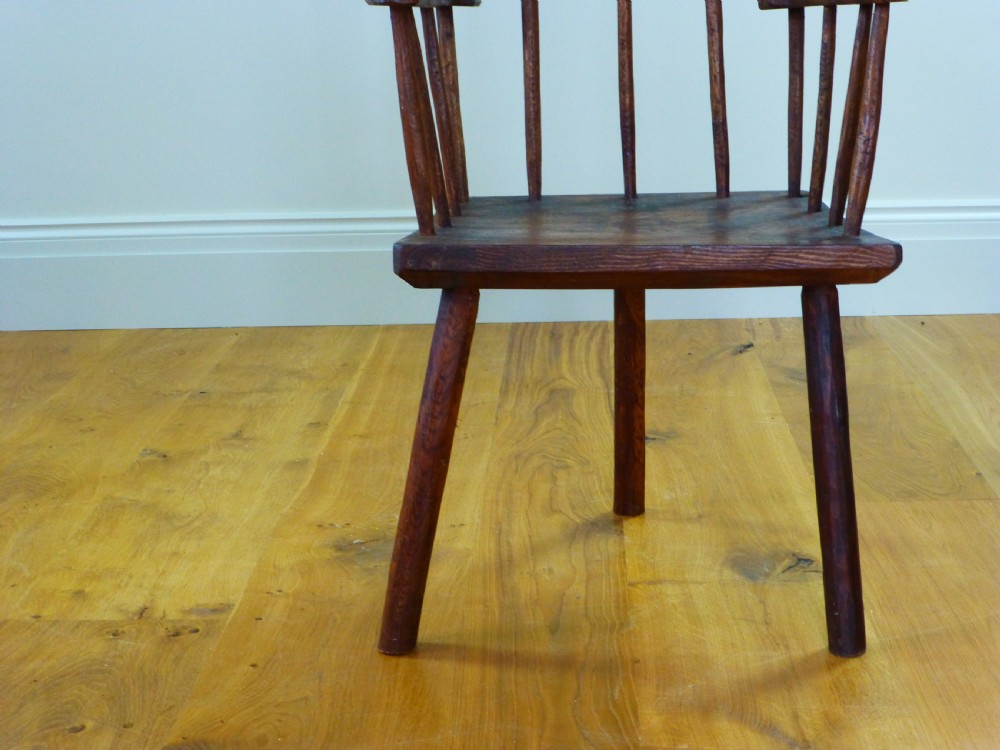 A primitive dark brown wooden Welsh stick armchair featuring a comb back crest rail, hand-shaped spindles, flat armrests, and a solid slab seat on splayed legs. View VII