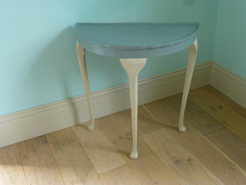 A painted demi lune console table featuring a light blue top surface and cream three-legged base with slender pad feet.