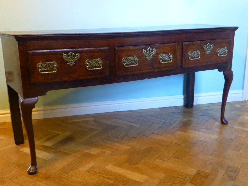 An 18th-century dark oak Georgian dresser base featuring three drawers with pierced brass handles and front cabriole legs.