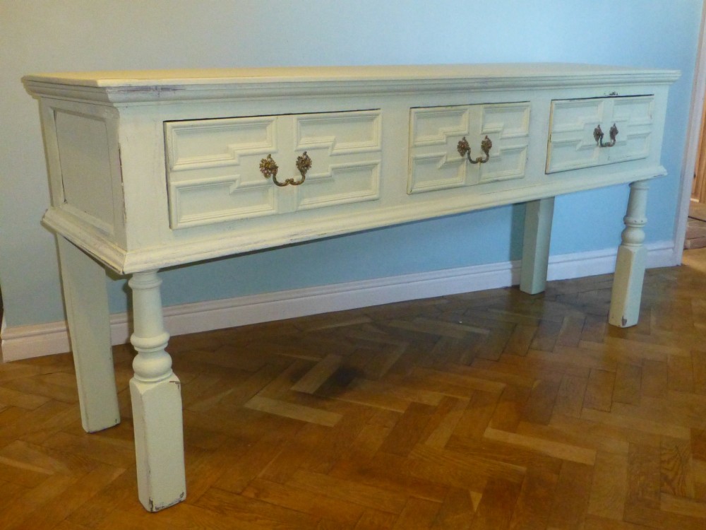 A long, cream-painted 17th-century style oak dresser base featuring three drawers with geometric mouldings, brass drop handles, and turned front legs.