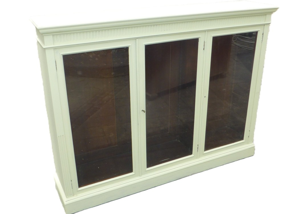A 19th-century off-white painted bookcase featuring three glazed glass doors, fluted side pilasters, and a contrasting dark interior.