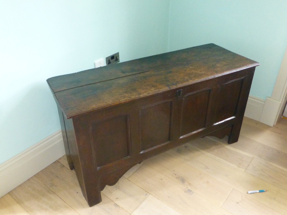 A dark oak 17th-century Welsh coffer featuring a four-panel front, hinged top lid, shaped lower apron, and integrated stile feet.