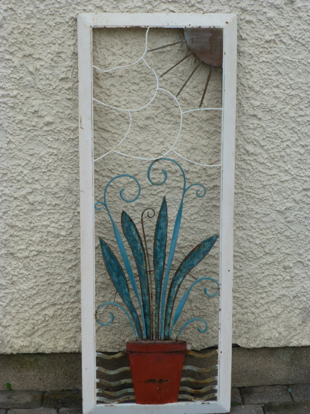 A rectangular white metal frame housing a decorative wrought iron panel with a sunburst motif and stylised blue potted plant design.