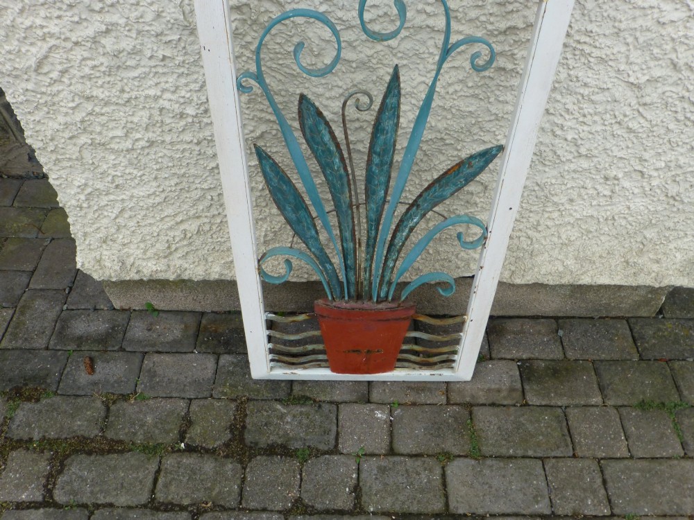 A rectangular white metal frame housing a decorative wrought iron panel with a sunburst motif and stylised blue potted plant design. View II