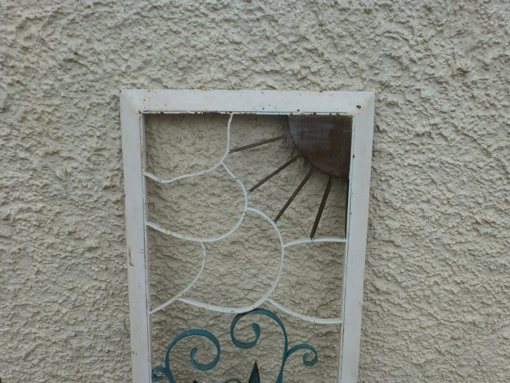 A rectangular white metal frame housing a decorative wrought iron panel with a sunburst motif and stylised blue potted plant design. View III