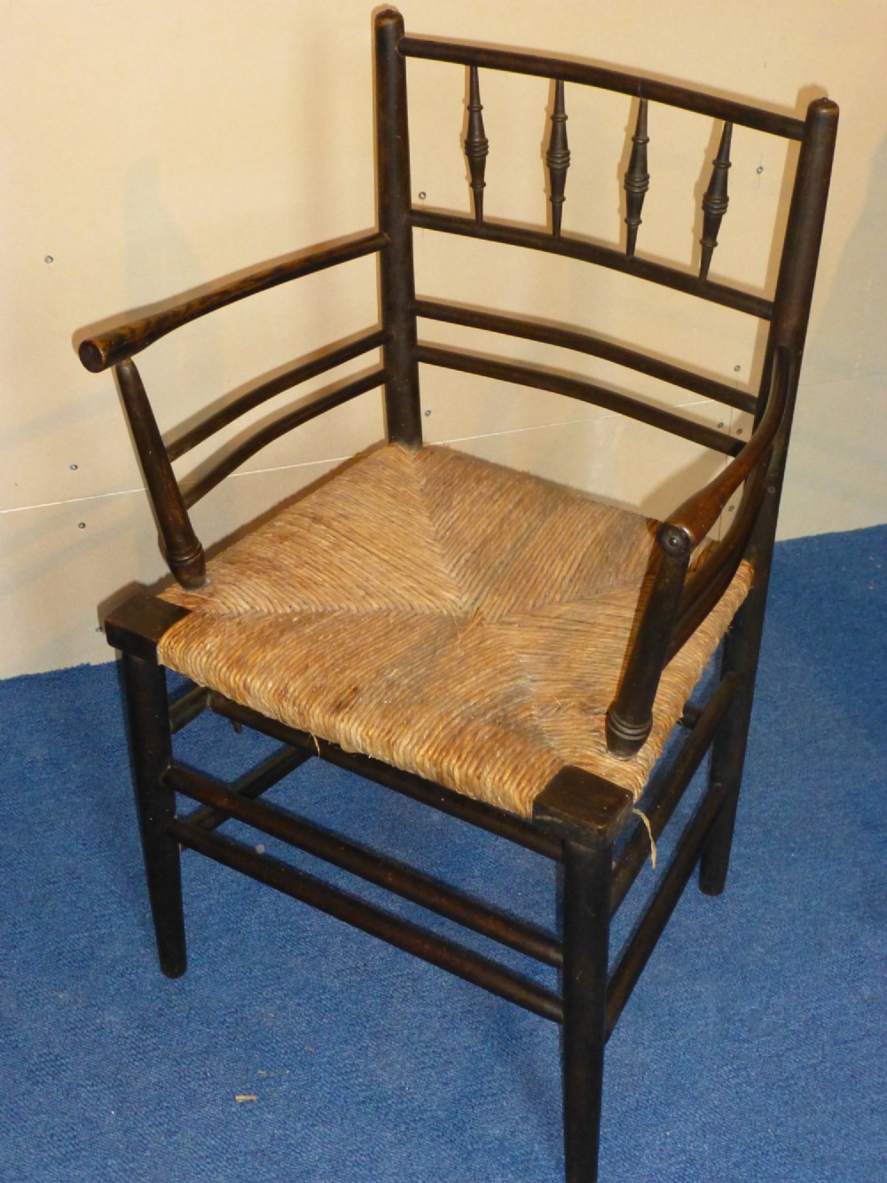 An Arts and Crafts ebonised wooden armchair featuring turned back spindles, curved armrests, and a woven rush seat. View II