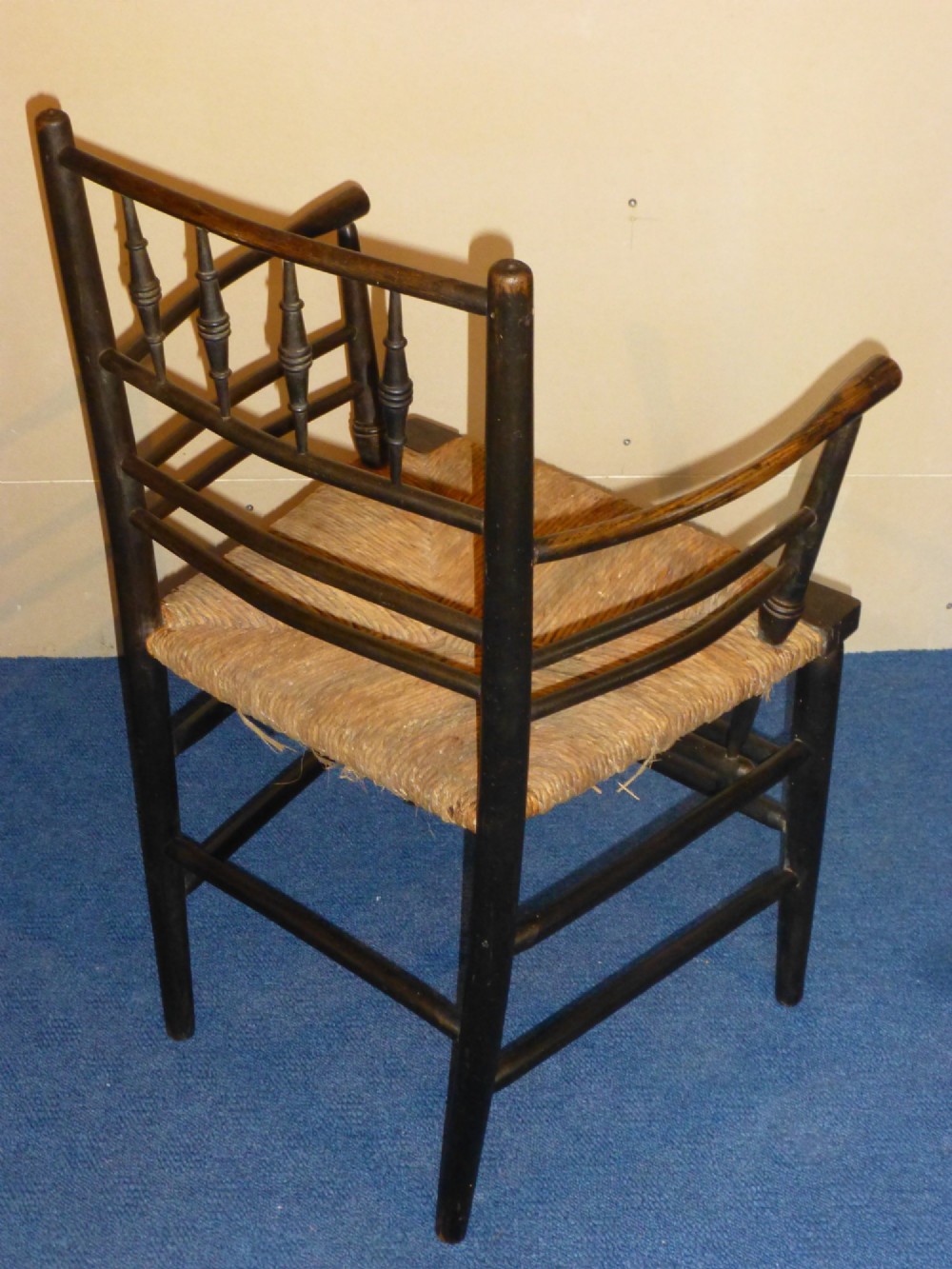 An Arts and Crafts ebonised wooden armchair featuring turned back spindles, curved armrests, and a woven rush seat. View III