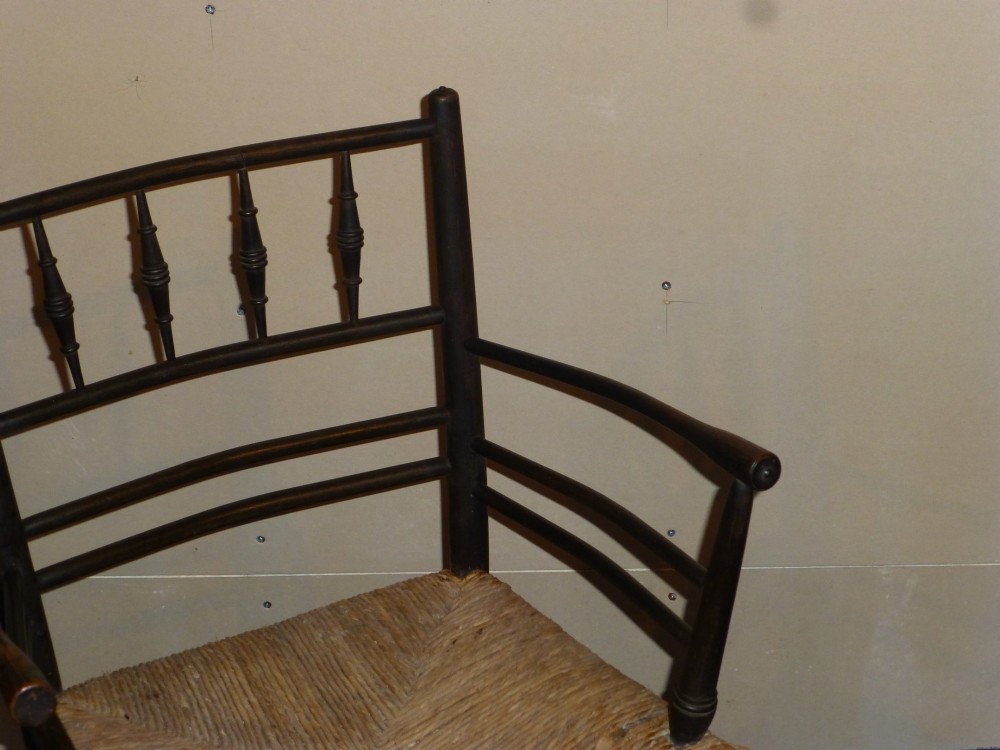 An Arts and Crafts ebonised wooden armchair featuring turned back spindles, curved armrests, and a woven rush seat. View V