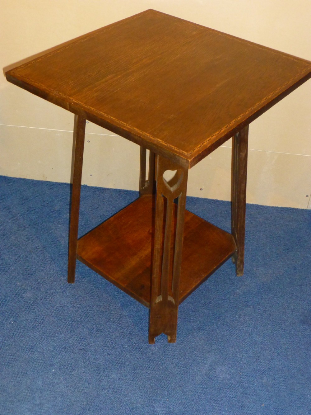 An Arts and Crafts solid oak occasional table featuring a square top with fine chequered inlay, a lower shelf, and vertical leg supports with heart-shaped cutouts.
