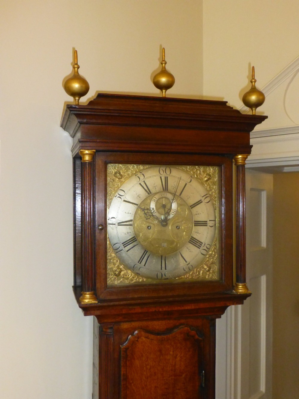 An eighteenth-century oak and walnut longcase clock featuring a square brass dial, decorative brass finials, and a slender veneered trunk door. View II