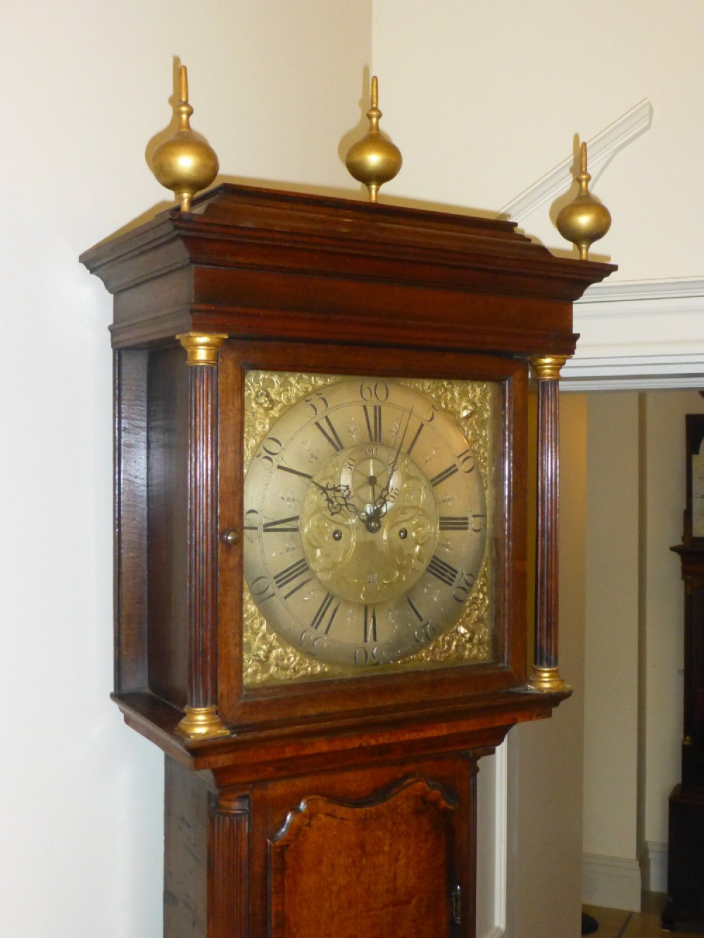An eighteenth-century oak and walnut longcase clock featuring a square brass dial, decorative brass finials, and a slender veneered trunk door. View IV