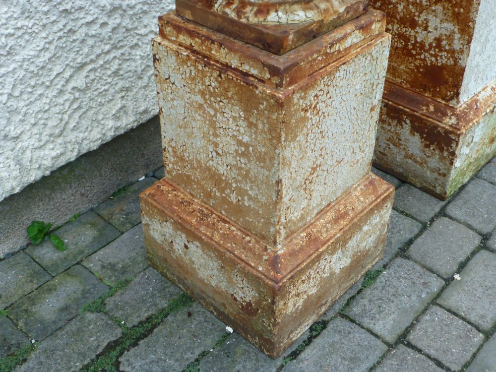A pair of Regency period cast iron campana urns with scrolled handles, displaying distressed off-white paint and surface rust, mounted on square cast iron pedestals. View IV