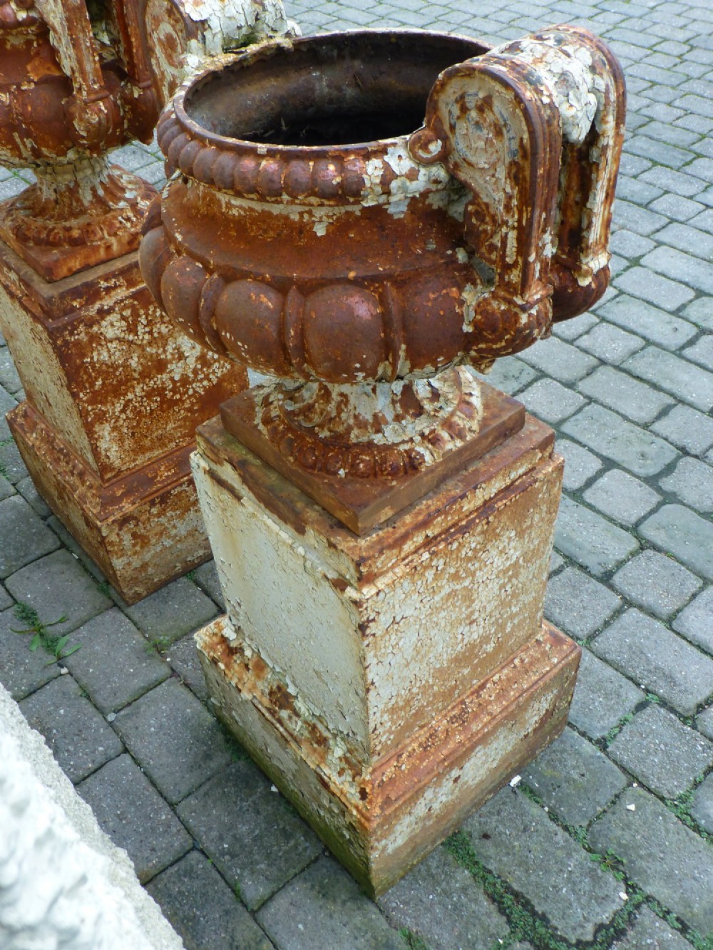 A pair of Regency period cast iron campana urns with scrolled handles, displaying distressed off-white paint and surface rust, mounted on square cast iron pedestals. View IX