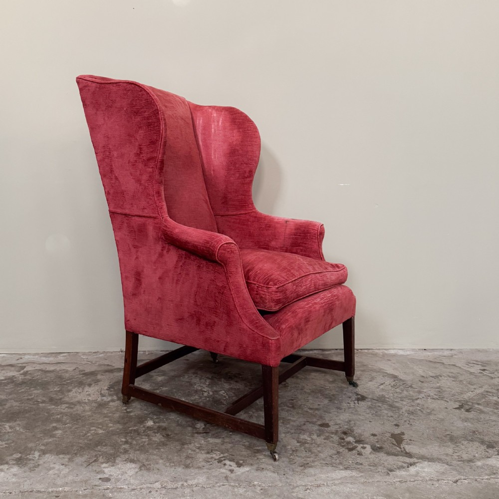 A classic English wingback armchair upholstered in red velvet with square timber legs, an H-stretcher base, and brass castors.