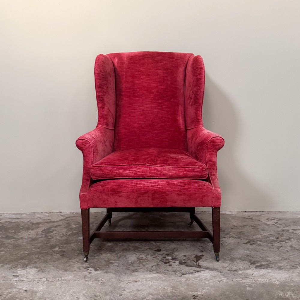 A classic English wingback armchair upholstered in red velvet with square timber legs, an H-stretcher base, and brass castors. View II