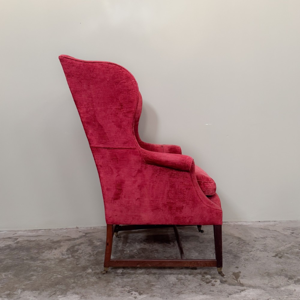 A classic English wingback armchair upholstered in red velvet with square timber legs, an H-stretcher base, and brass castors. View III
