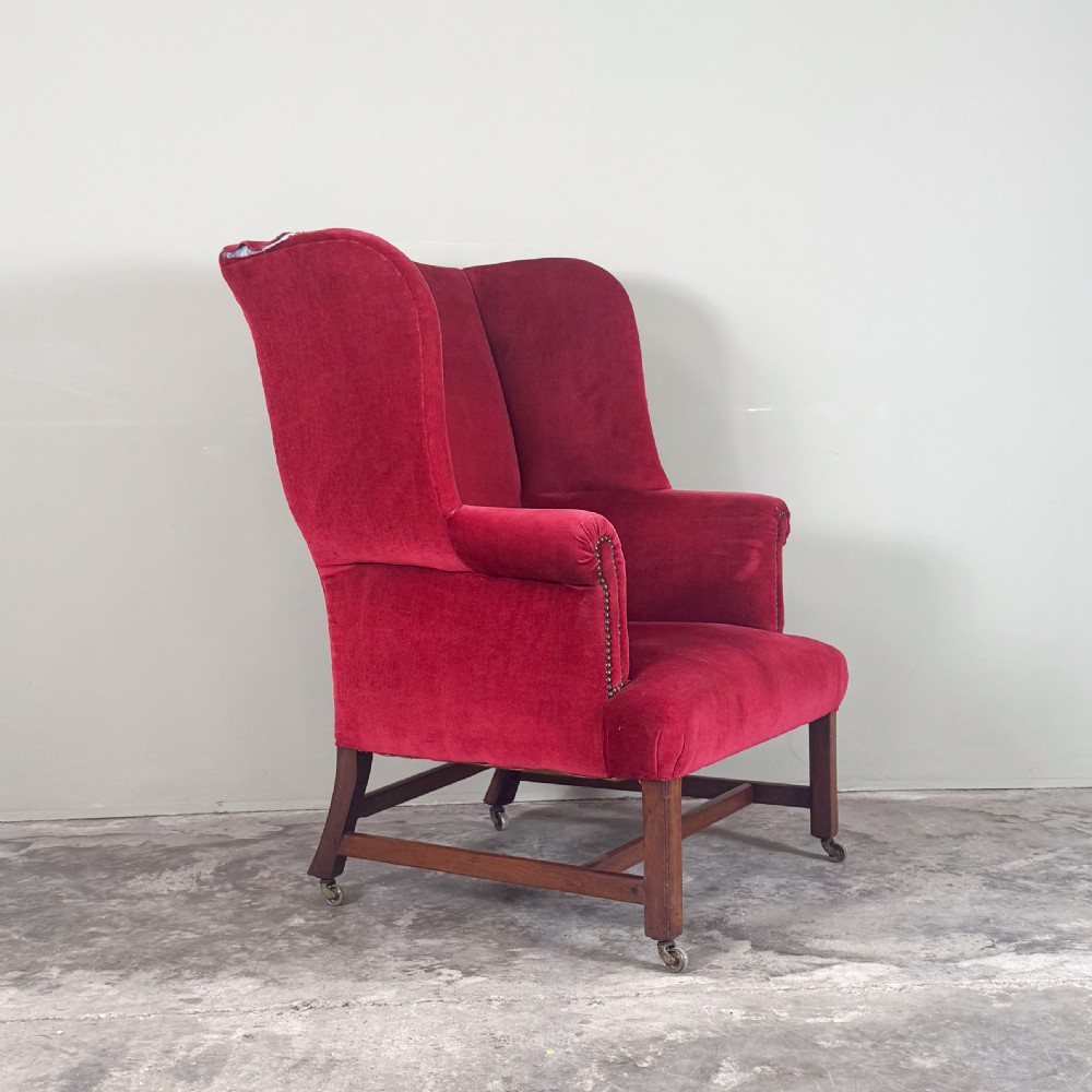 A 19th-century wingback armchair upholstered in red velvet with brass stud detailing on the arm fronts and timber legs on castors.