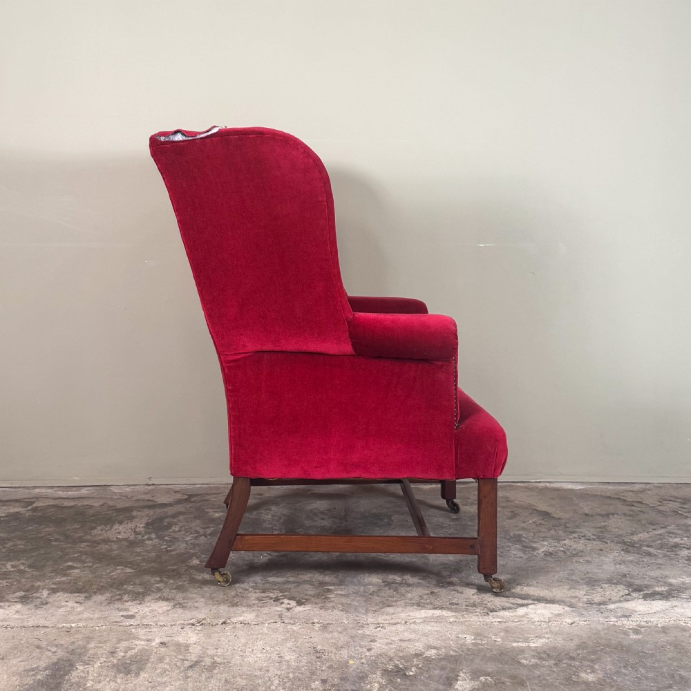A 19th-century wingback armchair upholstered in red velvet with brass stud detailing on the arm fronts and timber legs on castors. View II