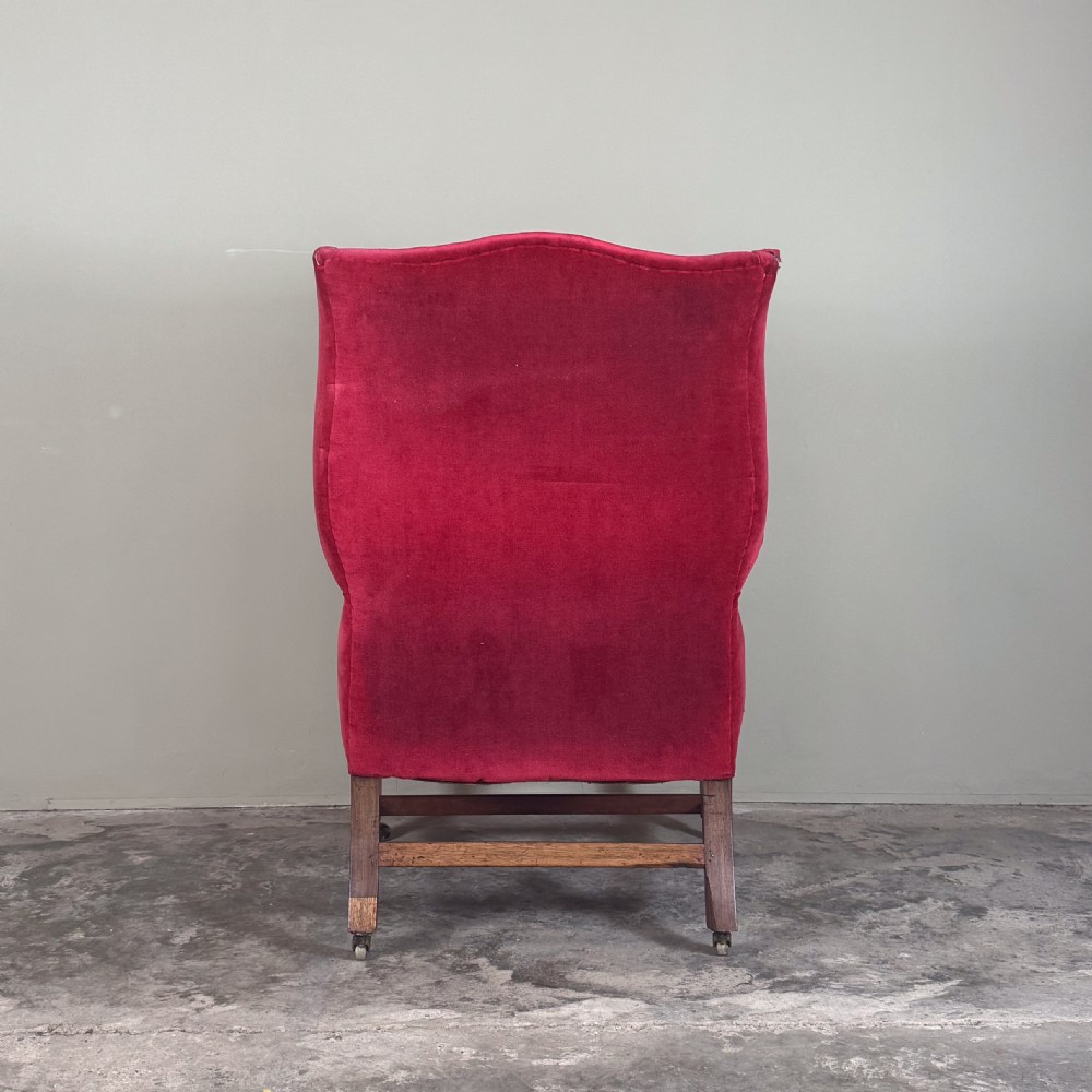 A 19th-century wingback armchair upholstered in red velvet with brass stud detailing on the arm fronts and timber legs on castors. View IV