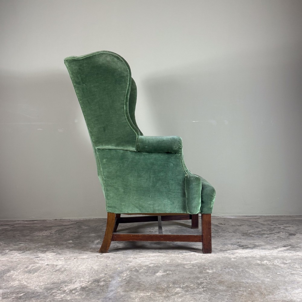 A high-backed antique wingback armchair upholstered in forest green velvet with dark wooden legs and stretchers. View III