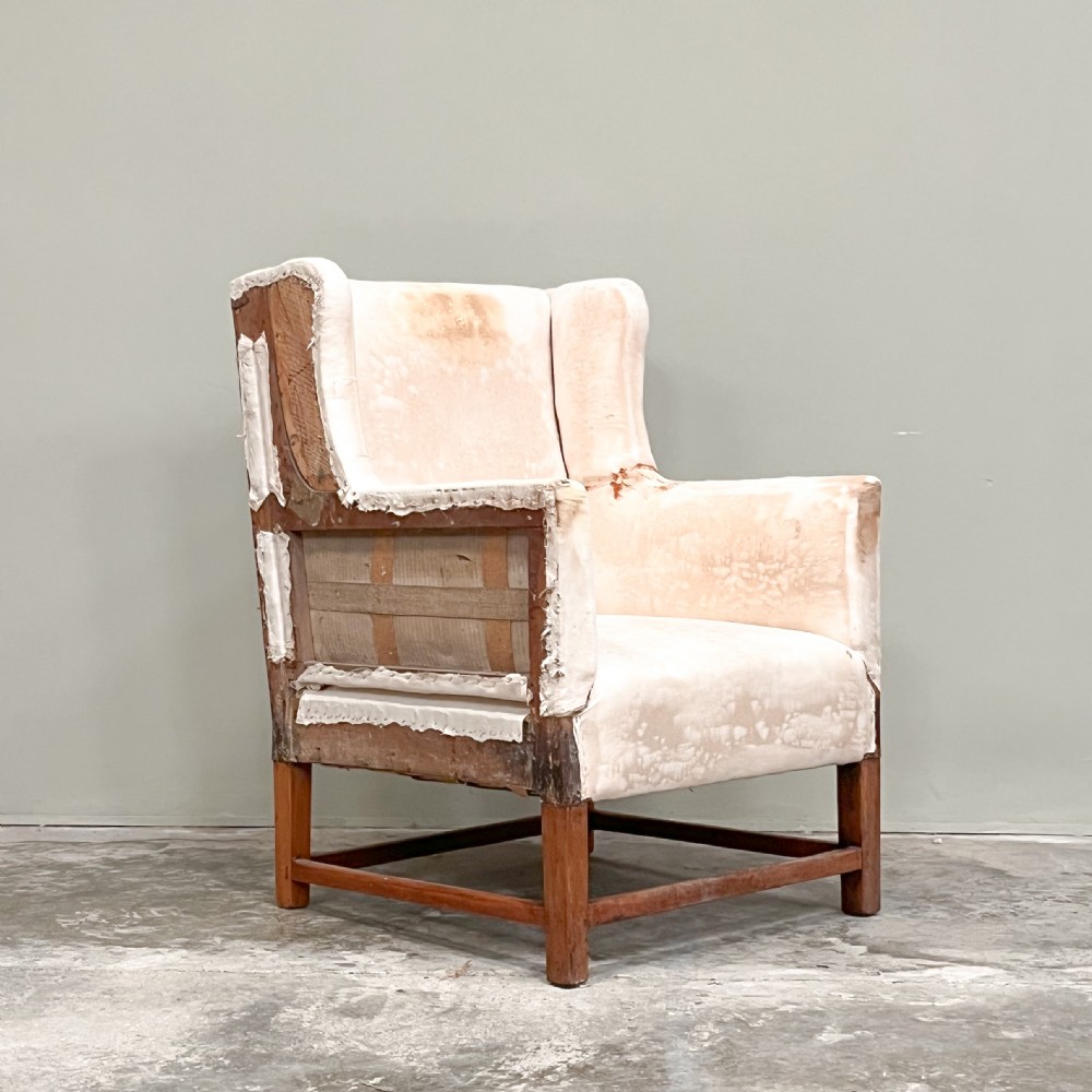 A 19th-century wingback armchair frame lined in cream calico fabric with exposed wooden side structure and square timber legs.