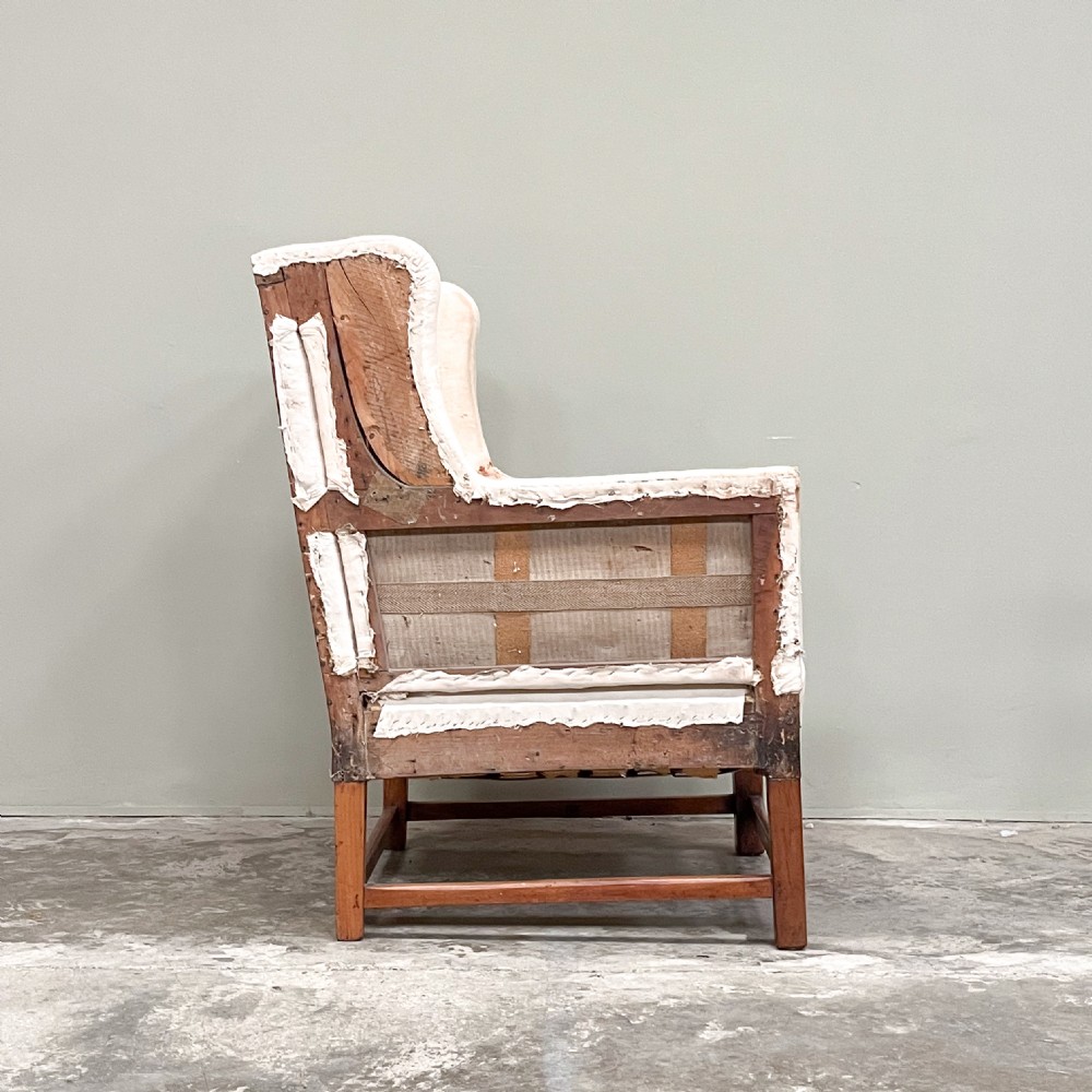 A 19th-century wingback armchair frame lined in cream calico fabric with exposed wooden side structure and square timber legs. View II