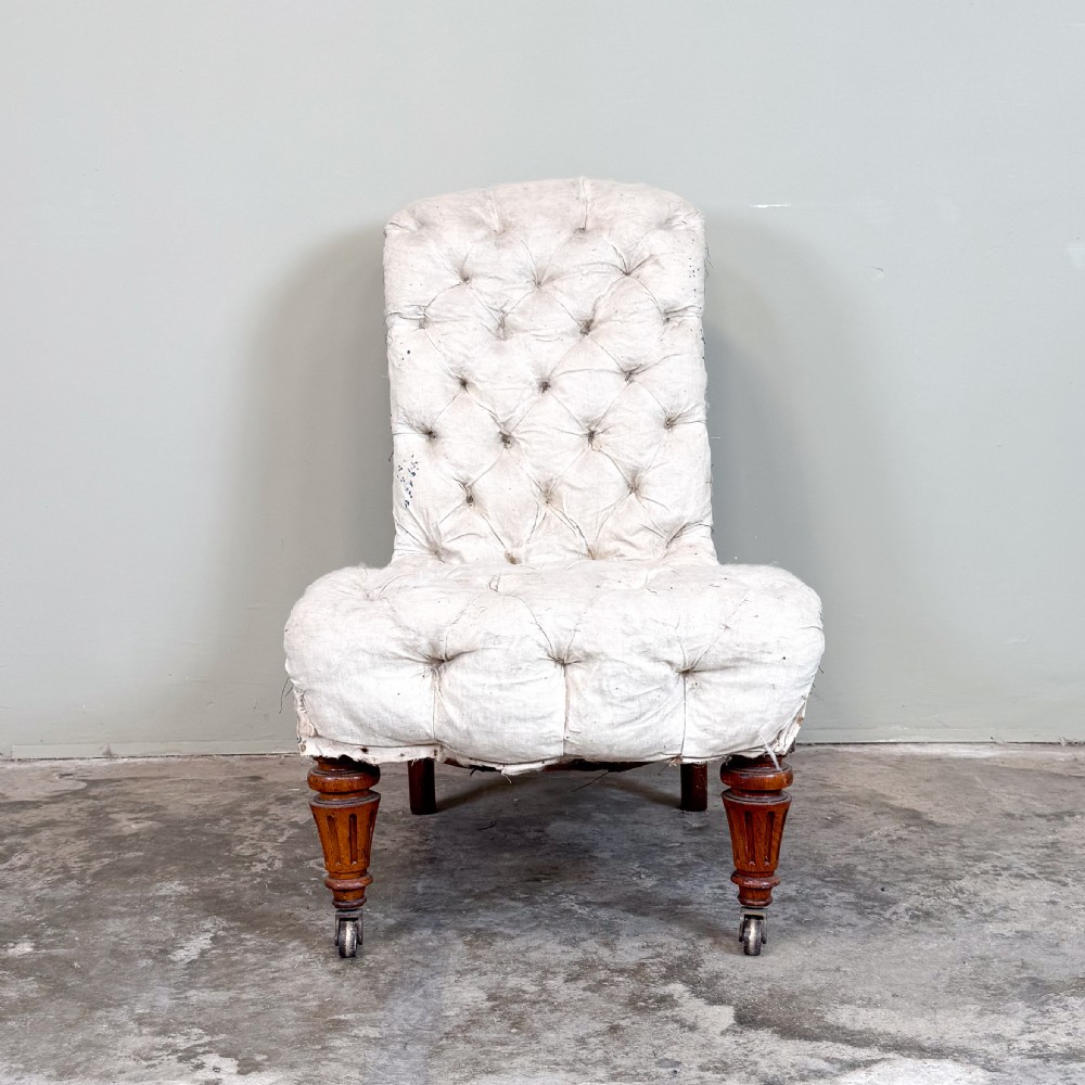 A Victorian armless slipper chair upholstered in off-white calico fabric with deep button-backed detailing and carved turned wooden legs on castors.