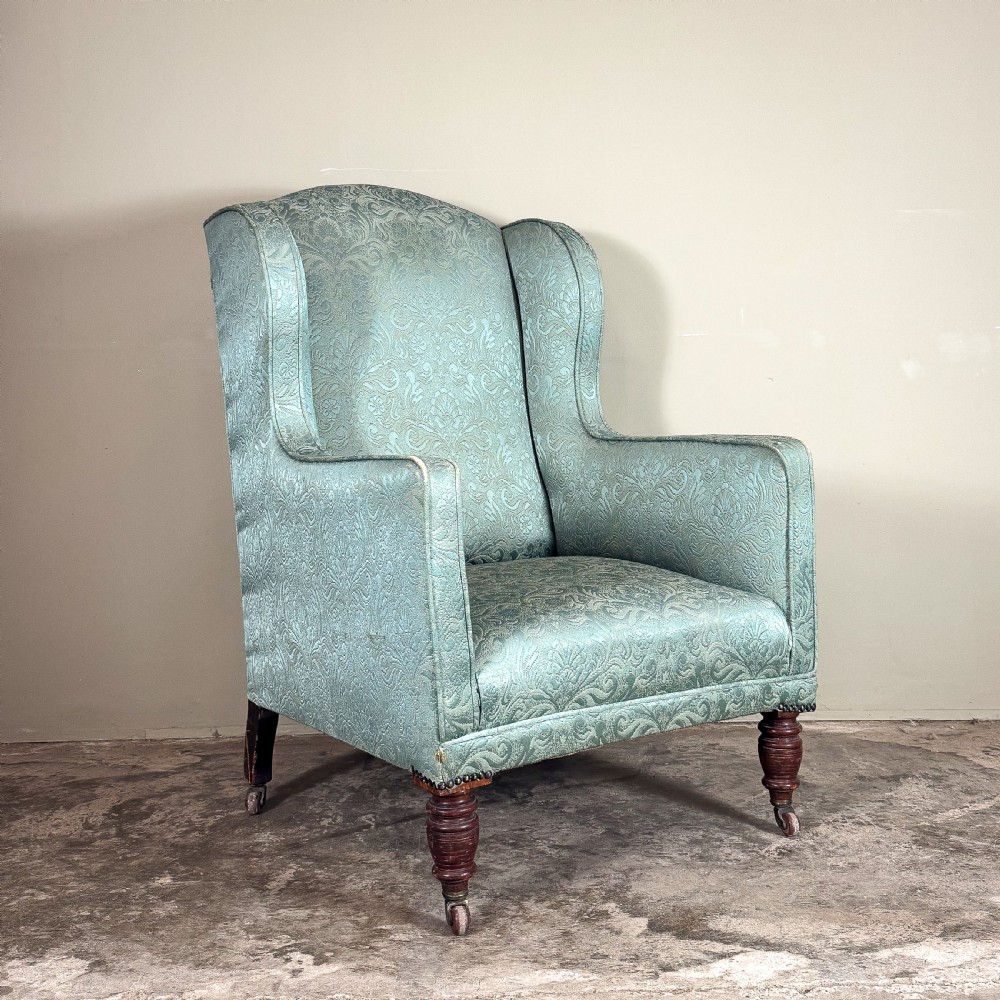 An Edwardian wingback armchair upholstered in duck egg blue patterned damask fabric with turned mahogany front legs on castors.