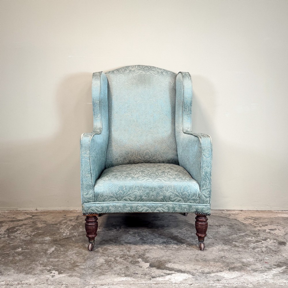 An Edwardian wingback armchair upholstered in duck egg blue patterned damask fabric with turned mahogany front legs on castors. View II