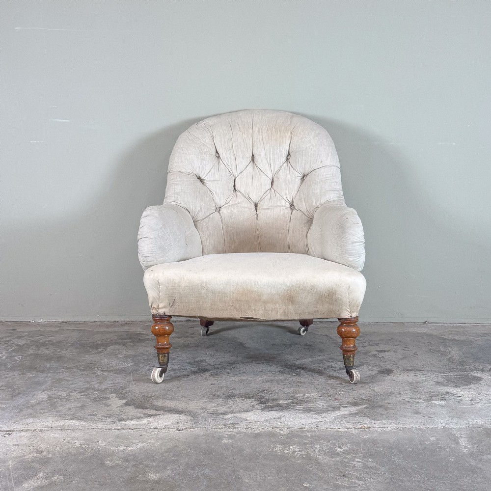 A 19th-century Victorian Howard style armchair upholstered in light cream fabric with deep button back tufting and turned wooden legs on brass castors.