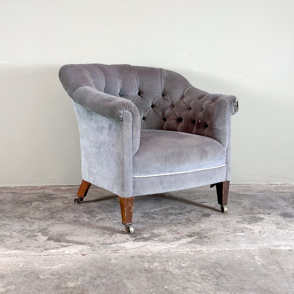 A Victorian-style grey velvet tub armchair featuring deep button tufting on the curved backrest, rolled arms, and tapered wooden legs on casters.