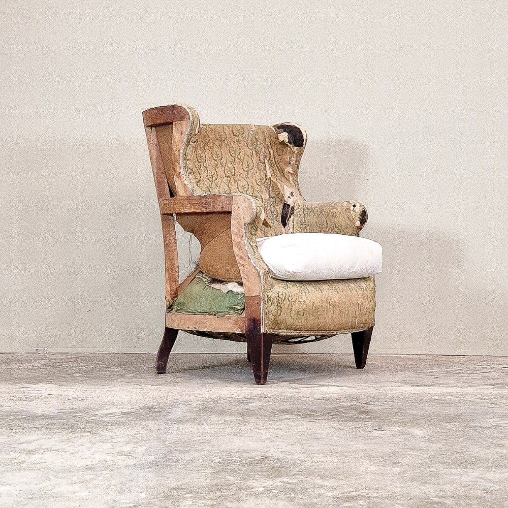 An Edwardian winged armchair shown in a partially deconstructed state with an exposed timber frame and worn green pattern upholstery.