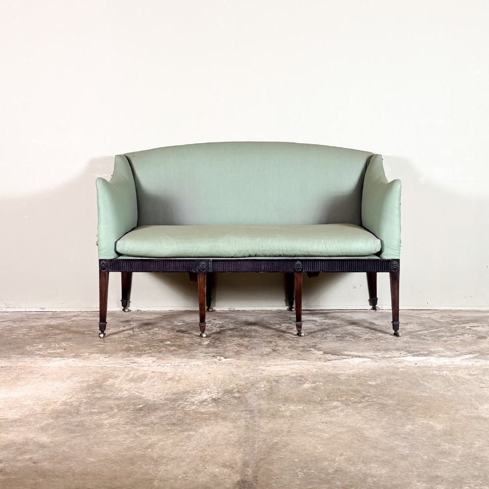 A Georgian two-seater sofa upholstered in light sage green fabric with a carved mahogany frame, fluted frieze, and tapered legs on brass castors.