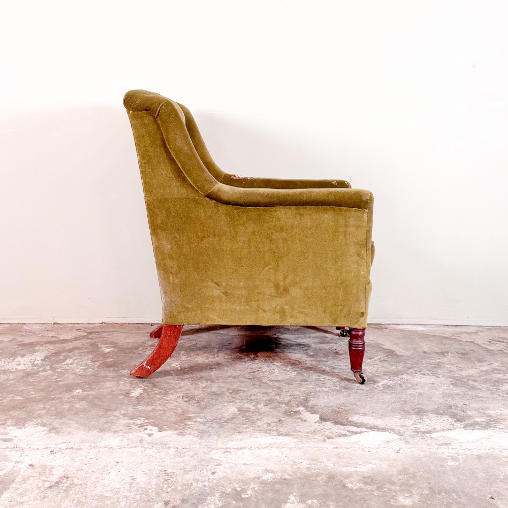 An antique Regency library armchair upholstered in olive green velvet with a buttoned backrest and turned mahogany front legs on brass castors. View II