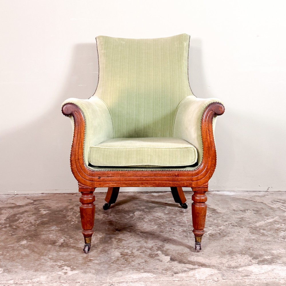 A Regency period library armchair upholstered in pale green velvet, featuring carved mahogany lyre-shaped arms and turned front legs on brass castors.