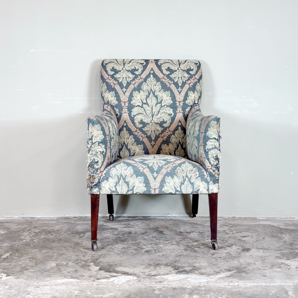 An Edwardian upholstered library armchair with damask patterned fabric in blue and pink, standing on tapered mahogany legs with brass castors.