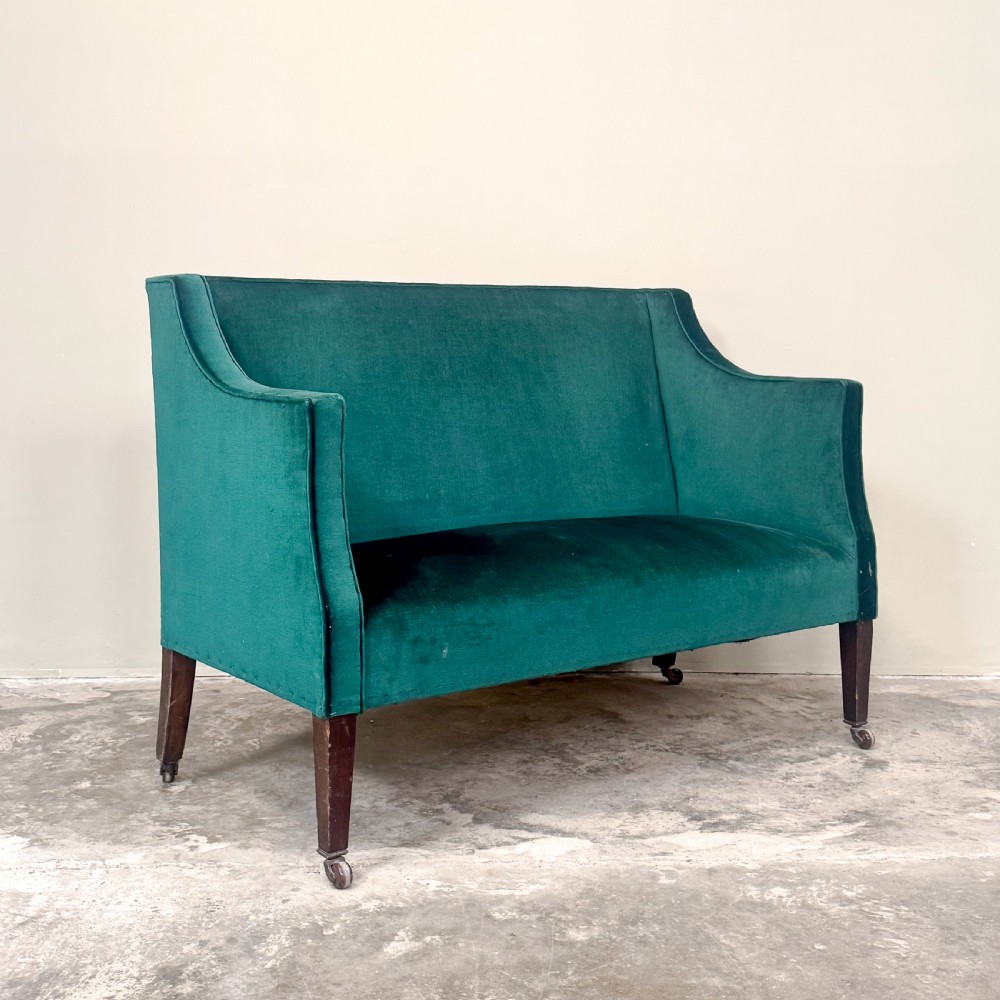 A petite Edwardian settee upholstered in emerald green velvet with sweeping sloped arms and square tapered mahogany legs on brass castors.