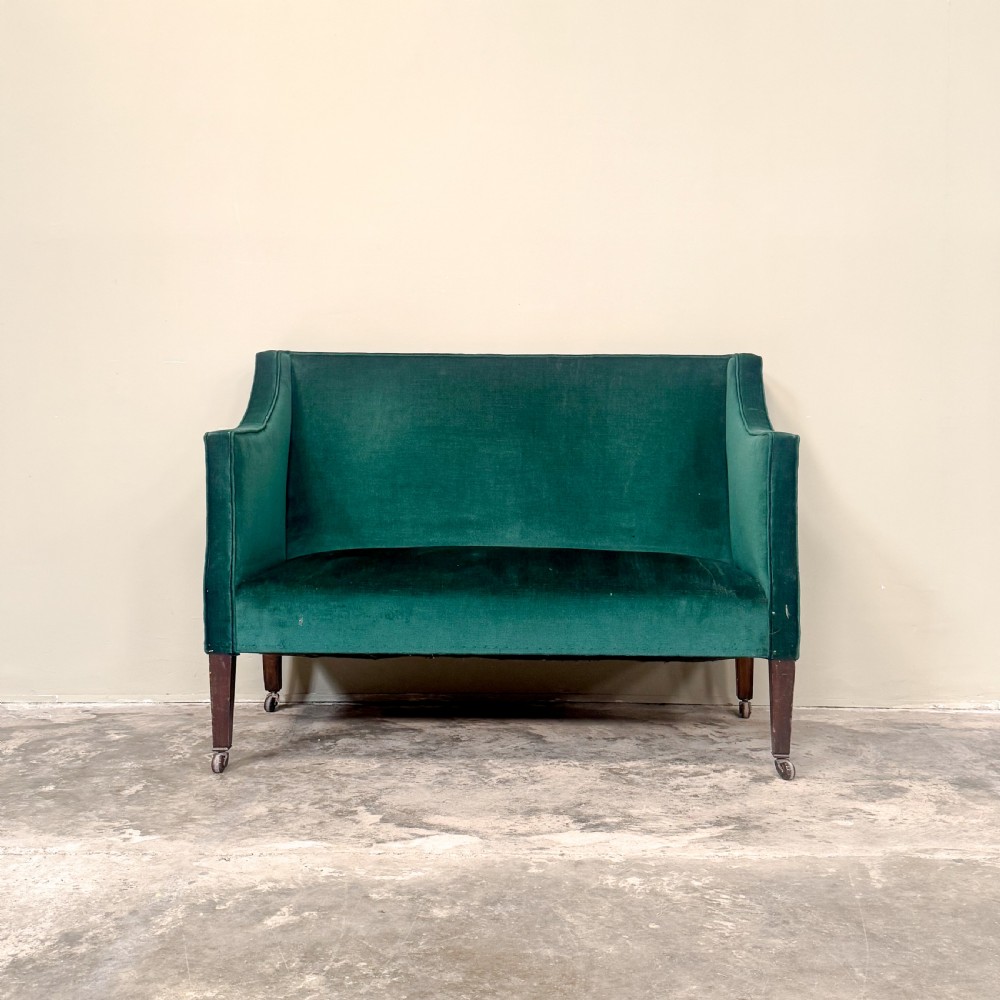 A petite Edwardian settee upholstered in emerald green velvet with sweeping sloped arms and square tapered mahogany legs on brass castors. View II