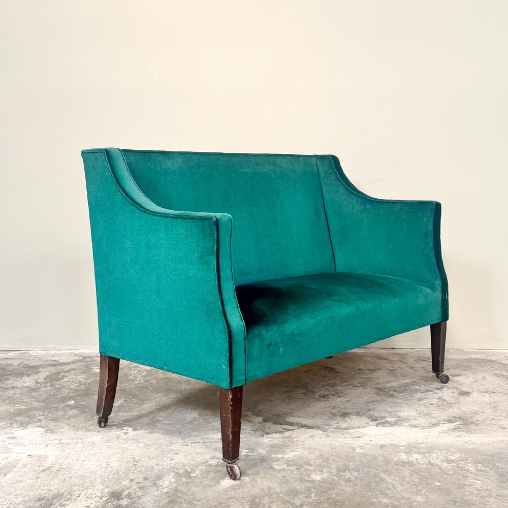 A petite Edwardian settee upholstered in emerald green velvet with sweeping sloped arms and square tapered mahogany legs on brass castors. View III