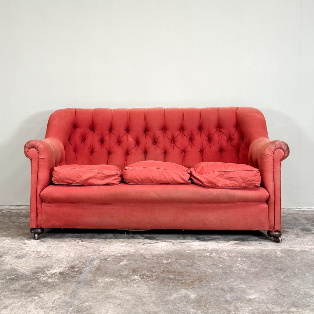A 1930s three-seater sofa featuring faded red upholstery with deep buttoning on the backrest, rolled arms, three loose seat cushions, and turned legs with castors. View II