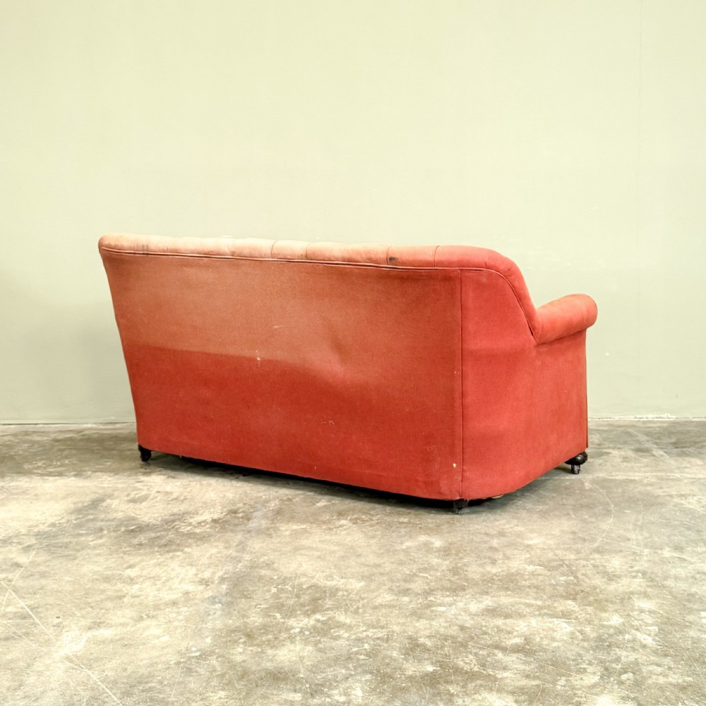 A 1930s three-seater sofa featuring faded red upholstery with deep buttoning on the backrest, rolled arms, three loose seat cushions, and turned legs with castors. View IV