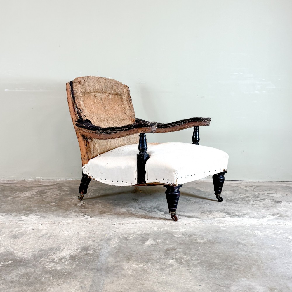 An antique open arm library chair stripped to its calico and hessian lining, featuring black turned front legs with brass castors.