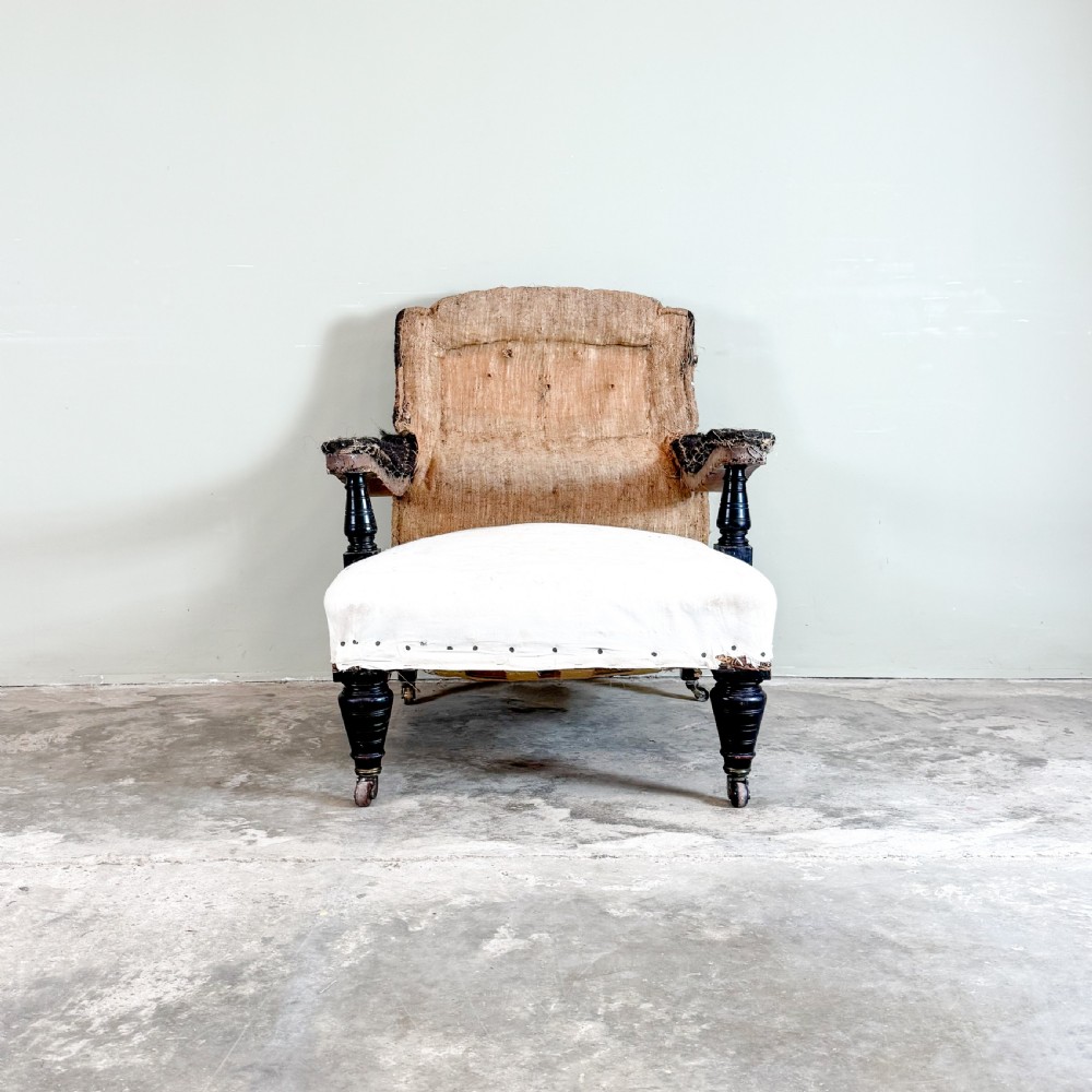 An antique open arm library chair stripped to its calico and hessian lining, featuring black turned front legs with brass castors. View III