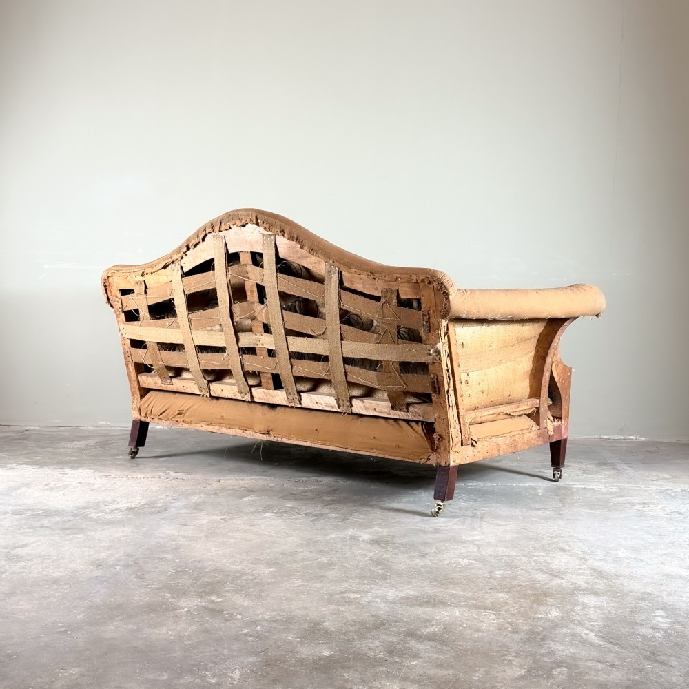 An Edwardian camel back sofa featuring a central arched backrest, exposed internal lining, loose seat cushions, tapered mahogany legs, and brass castors. View II