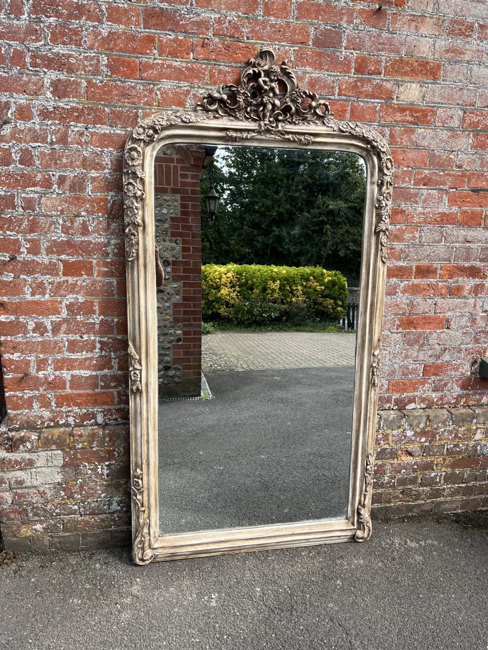 A large arched French mirror featuring an off-white painted wood and gesso frame decorated with floral carvings and a prominent cherub crest at the top. View IV